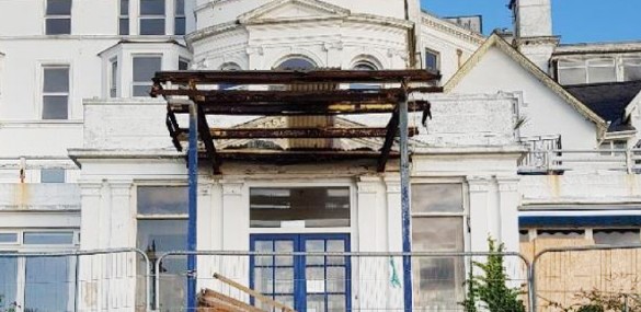Front façade of a dilapidated white building with boarded-up windows, blue double doors beneath a rusted metal canopy, and temporary railings in front.